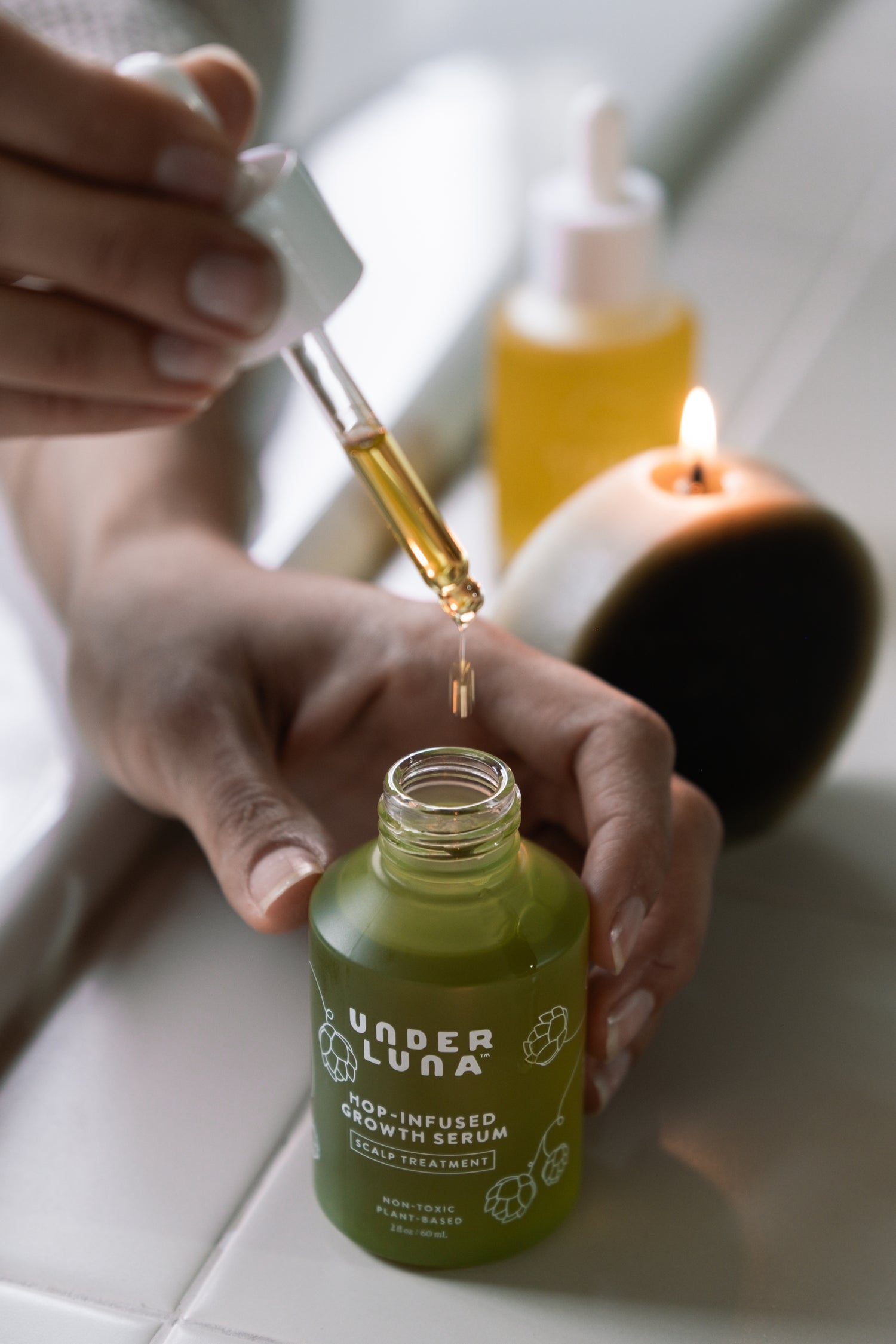 Hand holding a dropper over a green bottle labeled 'Under Luna' with a blurred candle in the background.