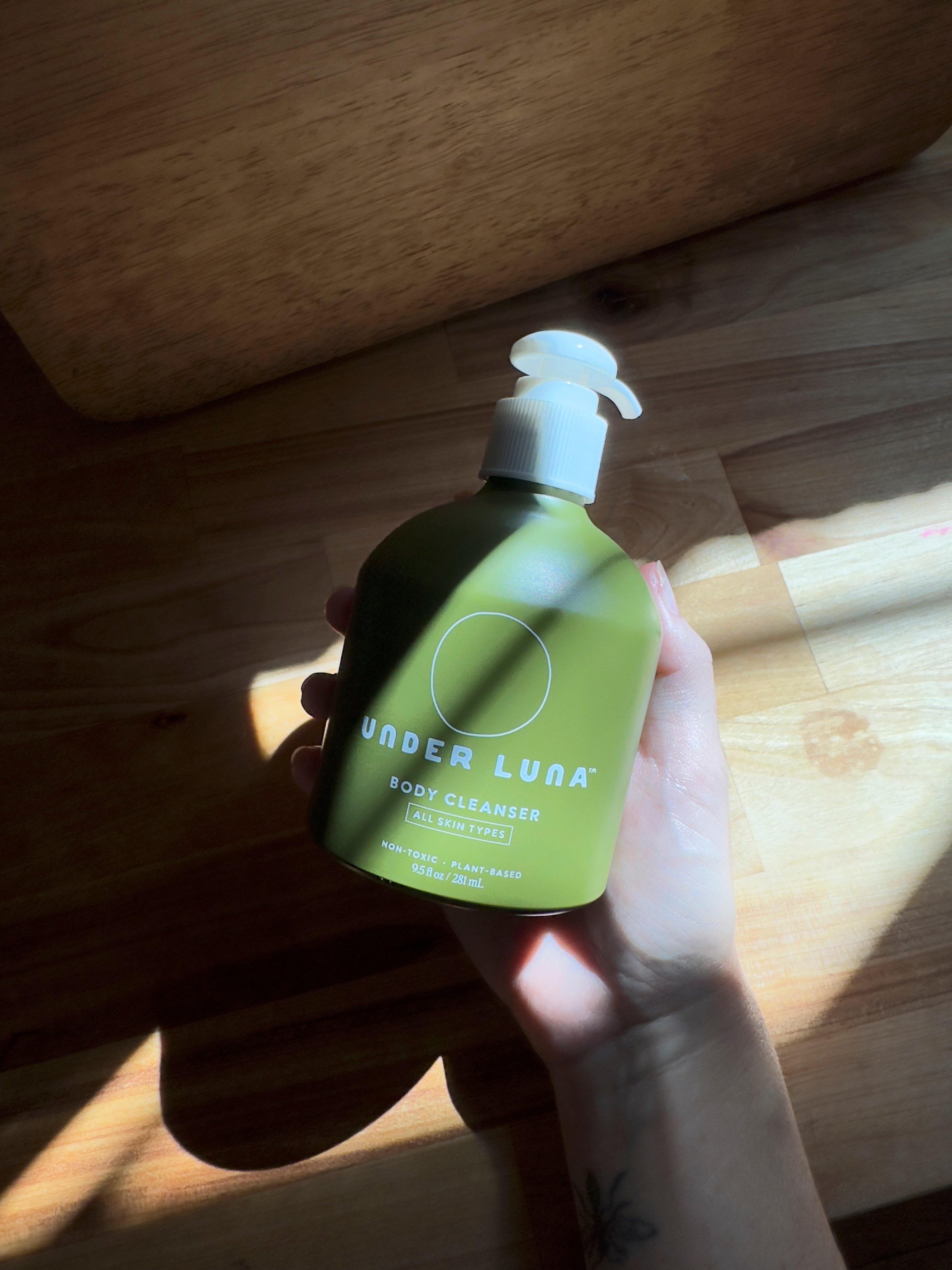 Hand holding a green bottle labeled 'Under Luna' on a wooden surface body cleanser wash shampoo holistic non toxic 
